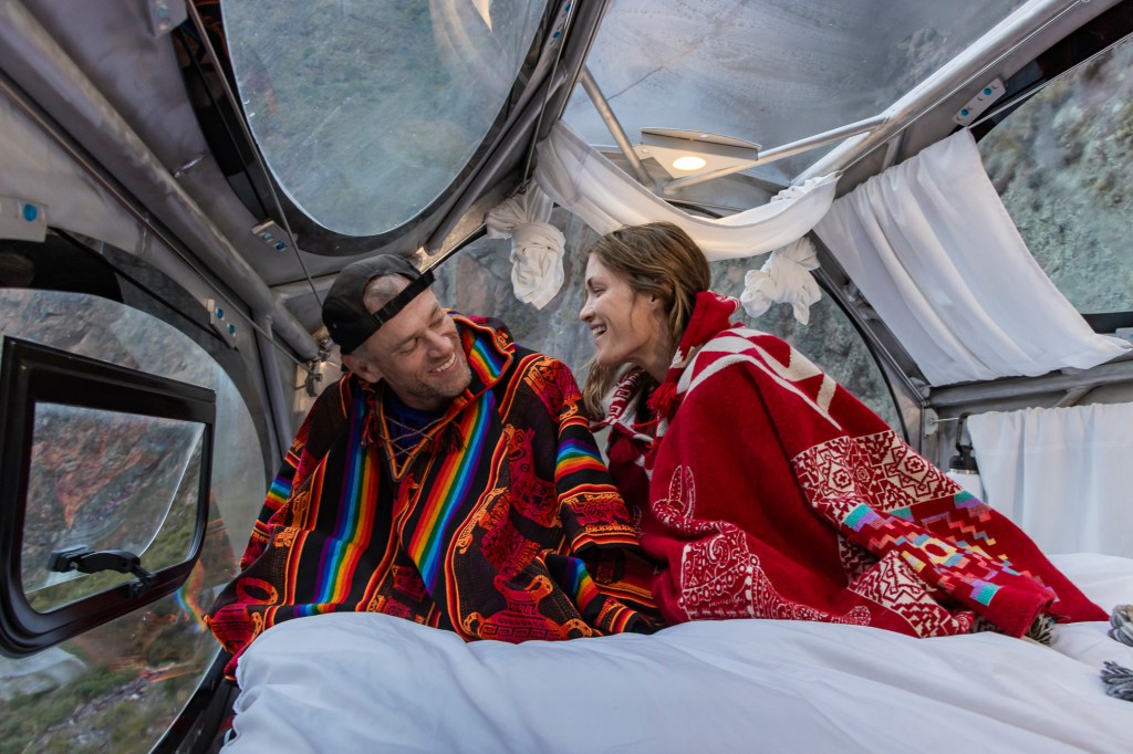 A couple enjoying a cozy moment inside a transparent capsule at Starlodge in Peru, wearing colorful ponchos and blankets with scenic mountain views outside.