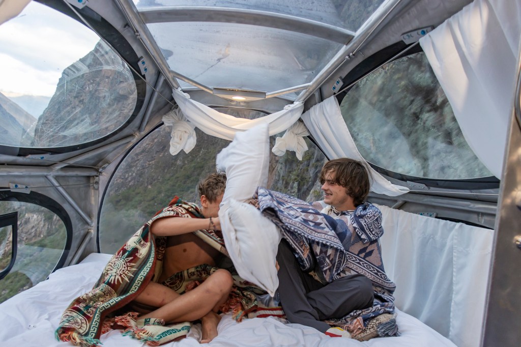 Two people playfully interacting inside a transparent capsule room with mountainous scenery in the background.
