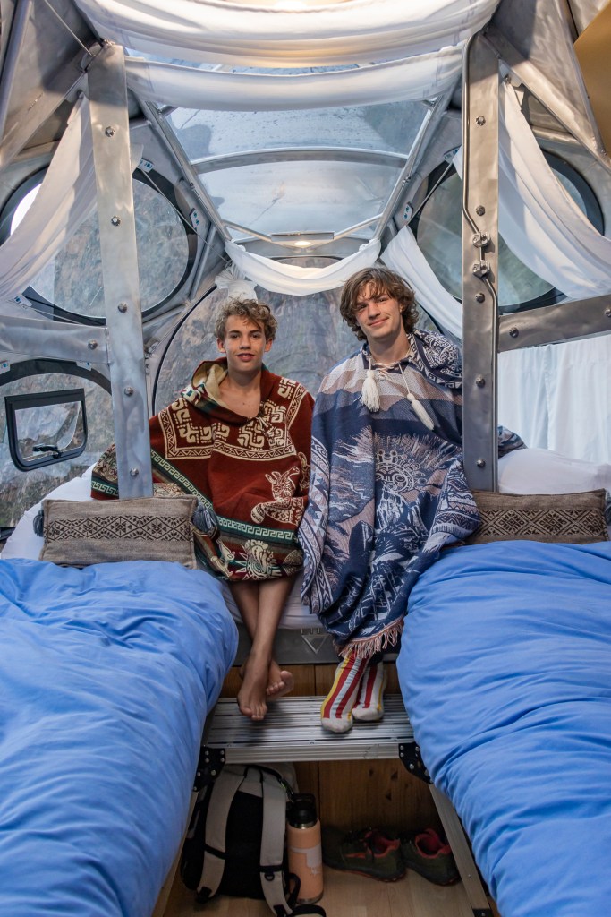 Two young men sitting on beds inside a transparent capsule-style hotel room, draped in colorful blankets with a scenic mountain backdrop visible through the windows.