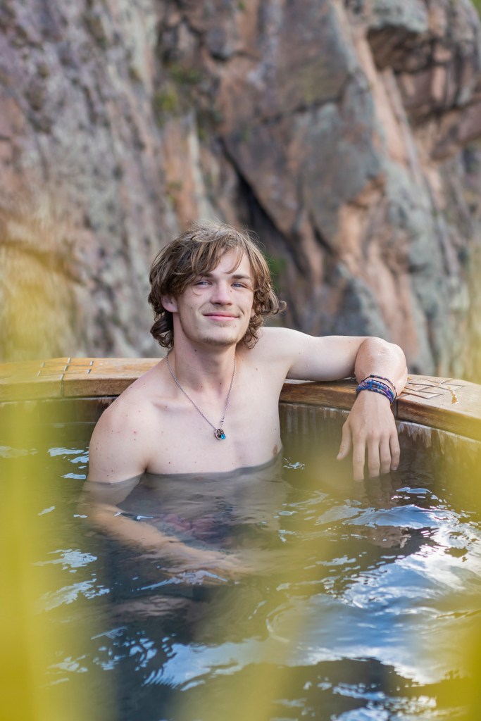A young man relaxing in a wooden hot tub surrounded by natural rock formations.