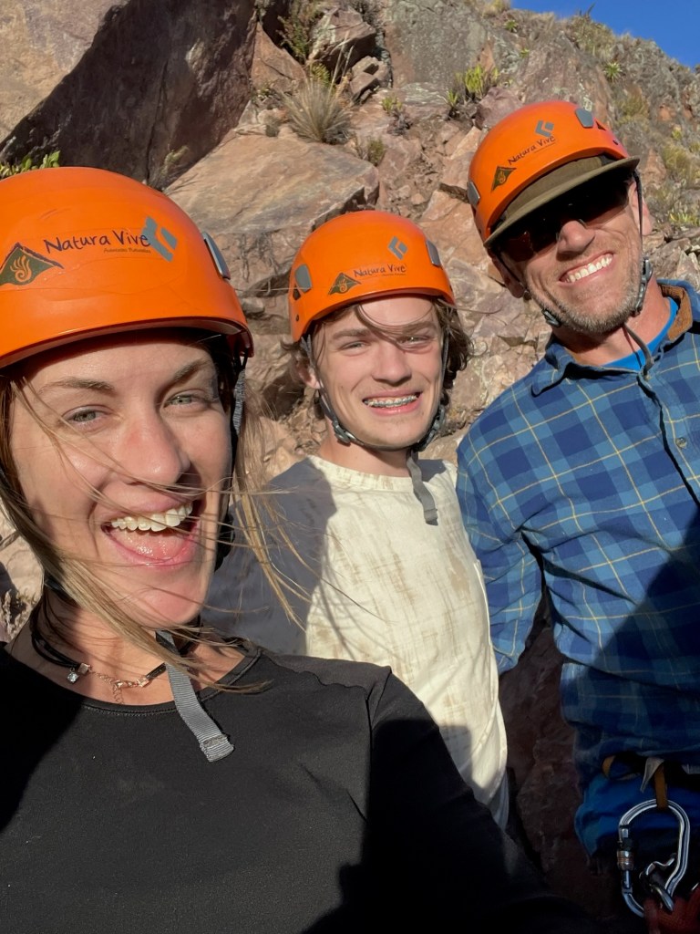 Three people smiling for a selfie while wearing orange climbing helmets, standing against a rocky cliff in a sunny outdoor setting.